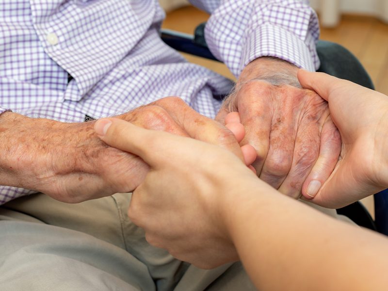 Close up detail of young female caregiver holding hands with old man in wheelchair.